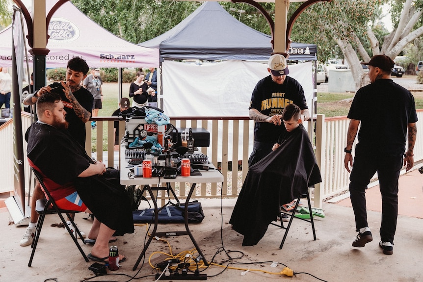 A mobile barber station has been set up in the park with two barbers giving haircuts to a man and a child.