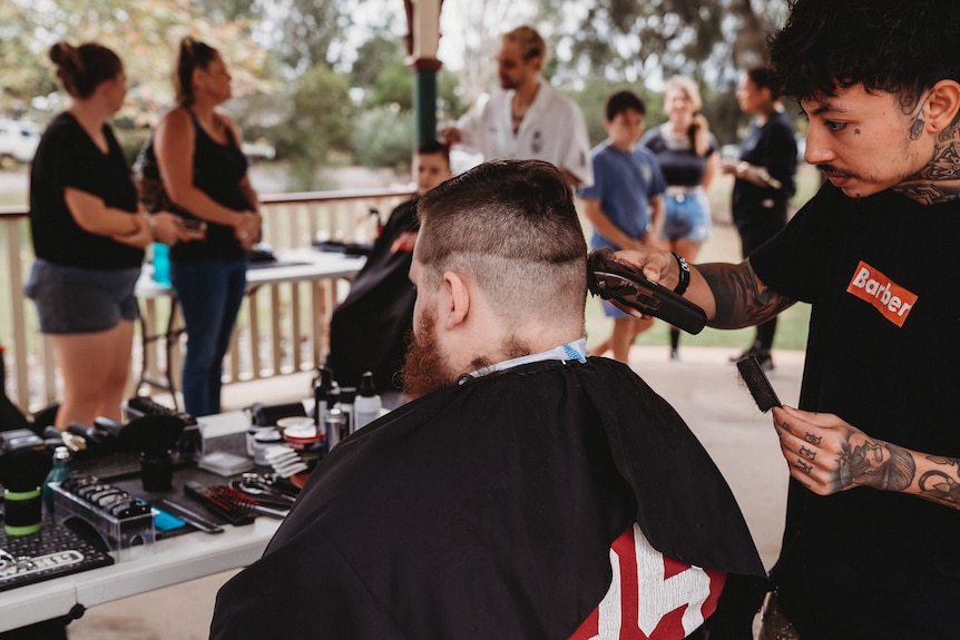 A male barber cutting hair cuts another man's hair in a park.