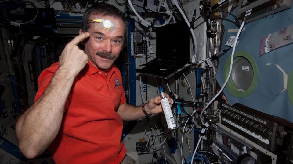 An astronaut points to a skin sensor on his forehead while floating in a space station module