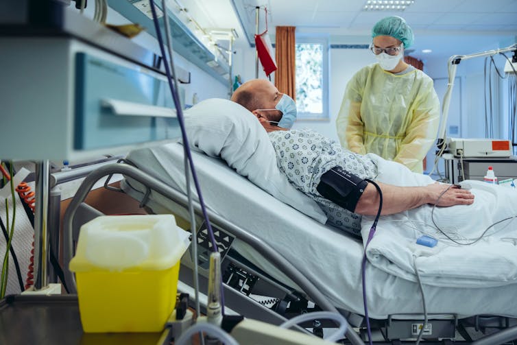 A woman in a surgical mask stands next to a patient in a hospital bed.