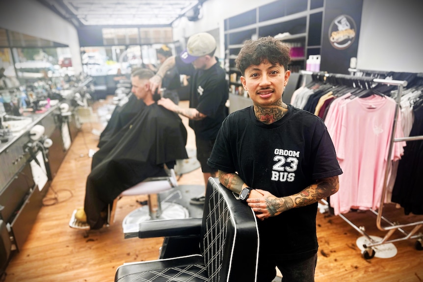 A male barber stands next to a barber's chair while other men cut hair behind.