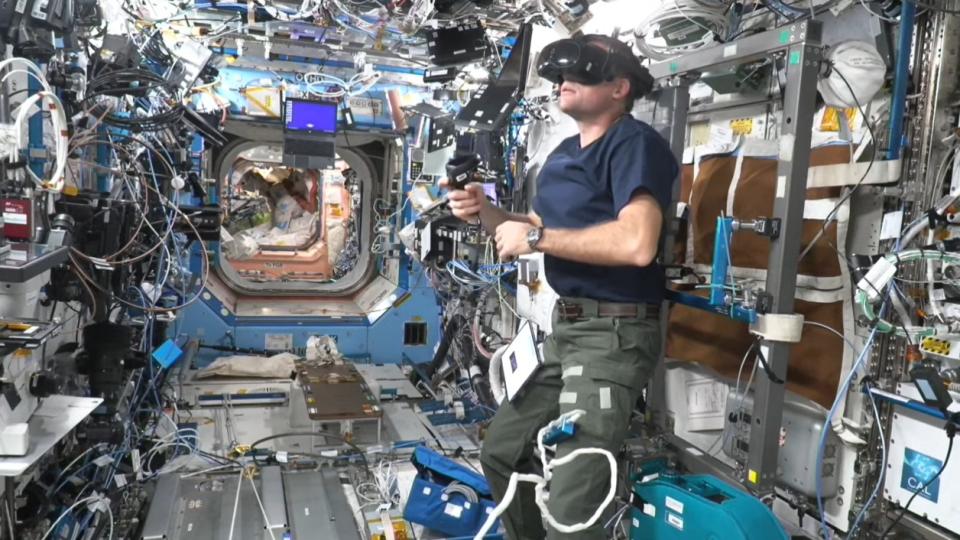 An astronaut wears a virtual reality headset in a crowded space station model.